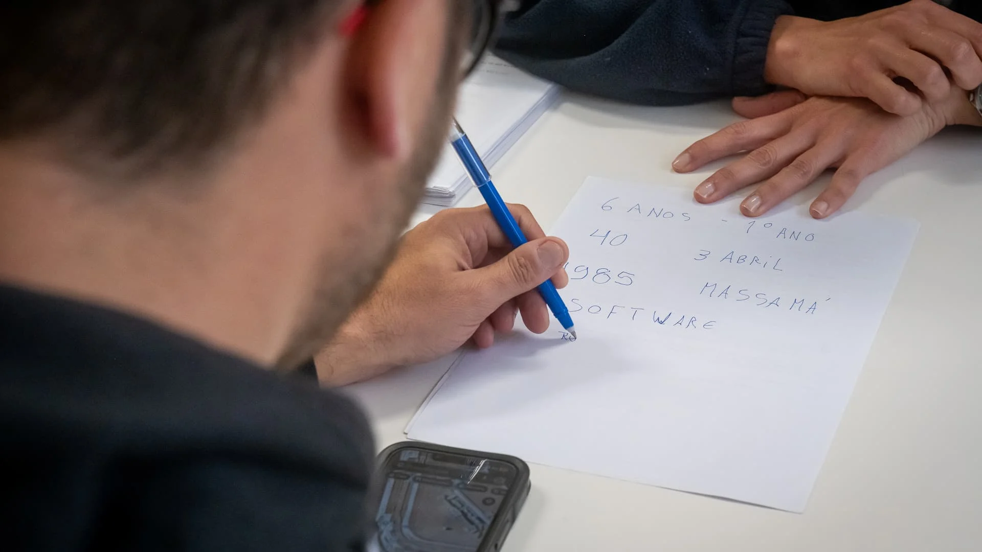 de costas, um homem escreve num papel branco algumas letras e números; à sua frente alguém observa mas apenas se veem as mãos dessa pessoas