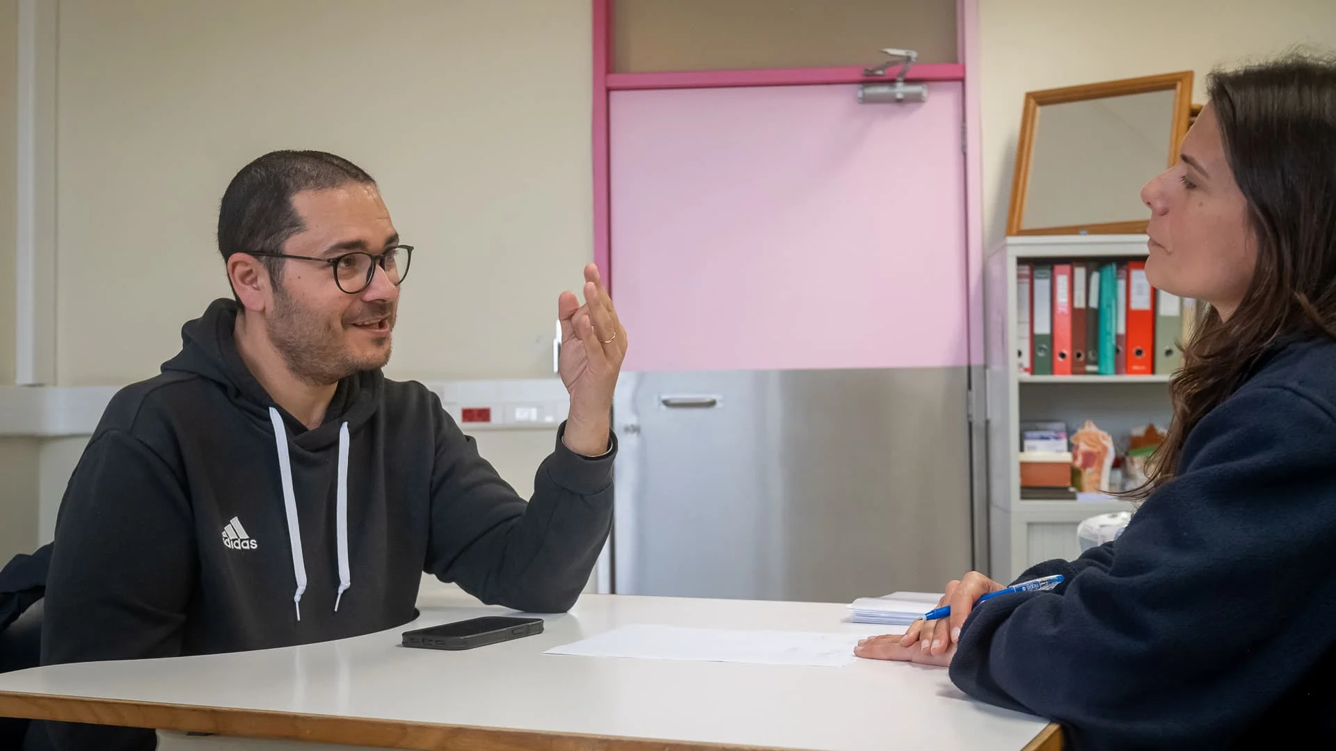 homem de óculos fala com uma pessoa que está à sua frente (mas cujo rosto não se vê); o homem fala com a mão esquerda levantada no ar