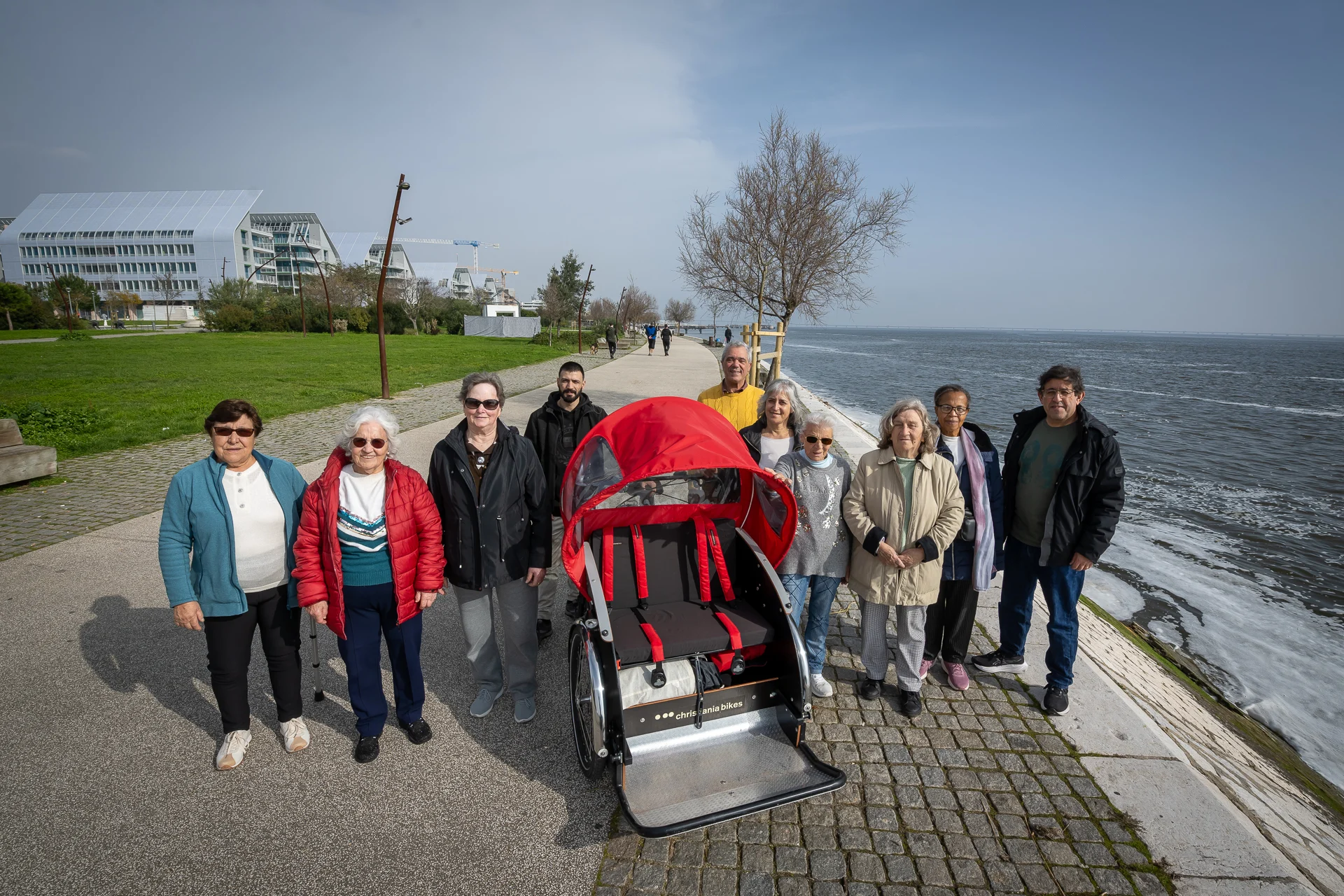 Capa_Passeio-Bicicleta Dez pessoas de pé, junto ao rio, posam para fotografia junto a bicicleta adaptada