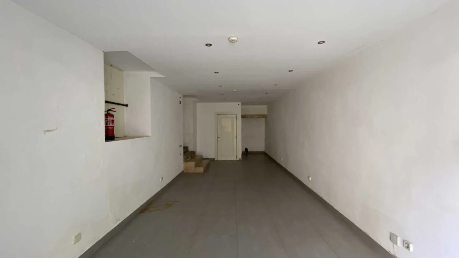 Long empty shop with a grey floor, a fire extinguisher on the wall and steps at the back
