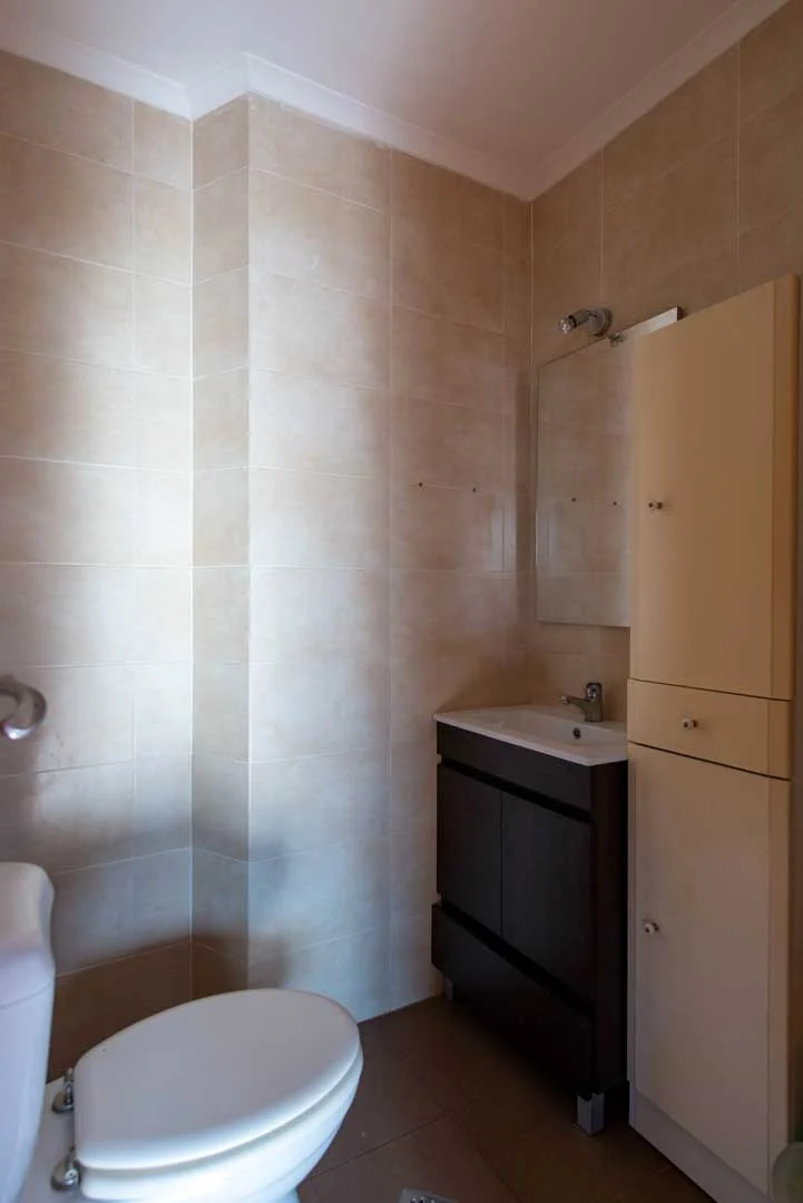 Casa de banho com azulejo bege, sanita, móvel de lavatório escuro e armário alto branco