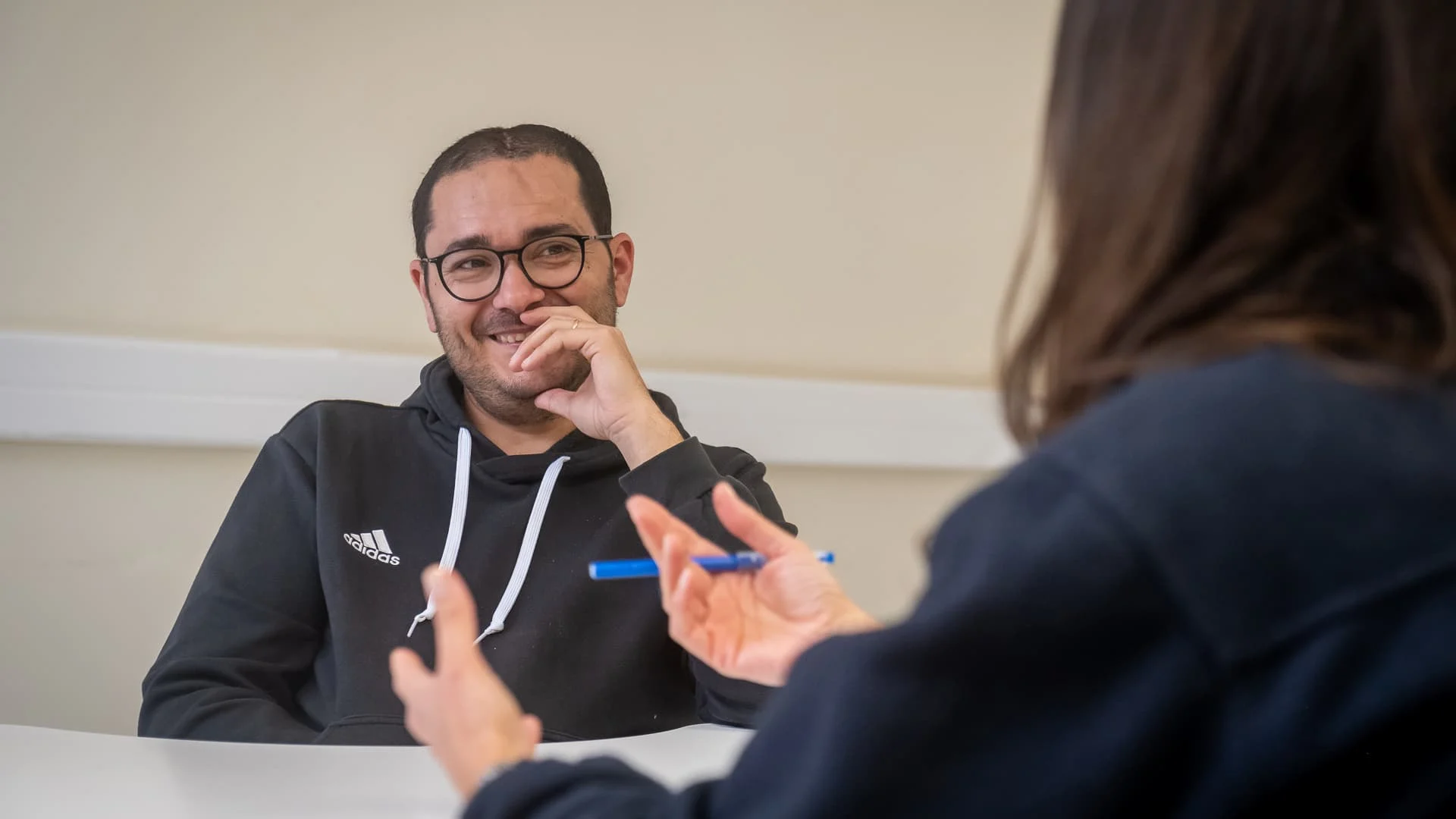 homem de óculos apoia mão no queixo expressando um sorriso timido para alguém com quem fala à sua frente (apenas se veem as mãos desta pessoa)
