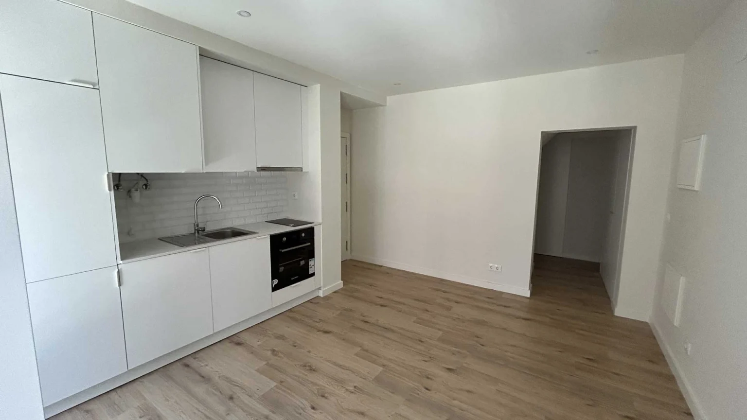 Sala com cozinha branca ao longo da parede, soalho claro e porta aberta para um corredor