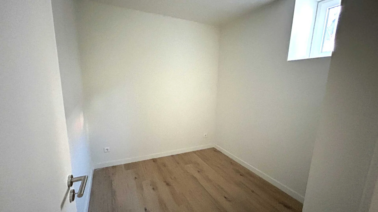 Quarto vazio de paredes brancas com janela alta e soalho claro