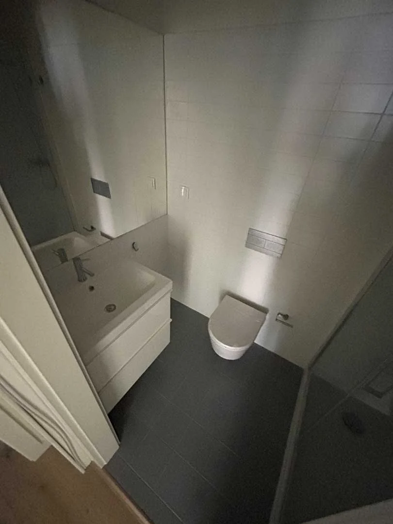 Casa de banho com chão escuro, sanita suspensa, lavatório branco e base de duche