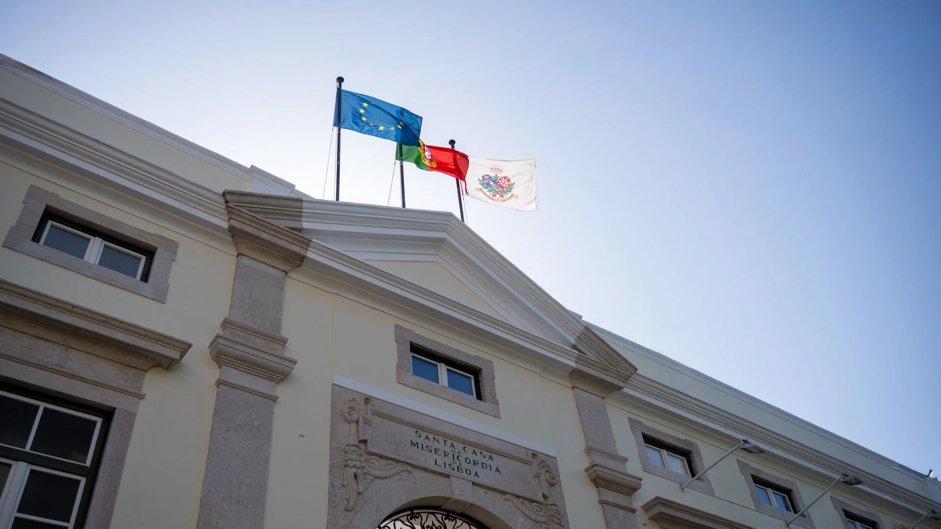 projetos-cofinanciados Fachada de entrada da santa casa da misericordia de lisboa com bandeiras da intituição, portugal e comunidade europeia